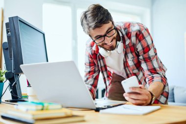 Gülümseyen programcı dizüstü bilgisayarda çalışıyor ve telefonu kontrol ediyor.