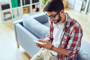 Evdeki mutlu genç adam telefonunu kanepeye dayayarak kullanıyor. 