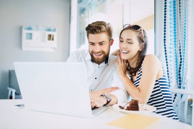Şık, parlak bir restoranda oturmuş laptopa bakıp gülen ve konuşan neşeli genç çift.