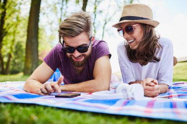 Genç çift parkta piknik yaparken battaniyeye uzanırken telefon kullanıyor.