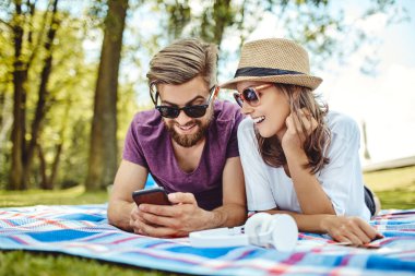 Parkta battaniyeye uzanmış akıllı telefon kullanan gülümseyen bir çift.