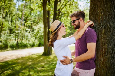 Genç çift parkta bir ağaca sarılıp yaslanıyor.