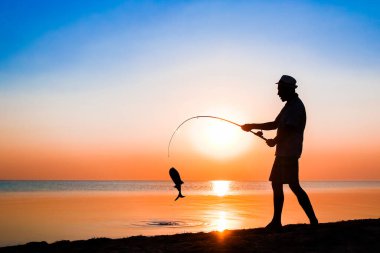 Mutlu balıkçı doğa siluetinde deniz kenarında balık yakalıyor.