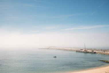 A view to Pescadores beach and pier with a little fishing boat in the ocean on a foggy day, Portugal, Ericeira