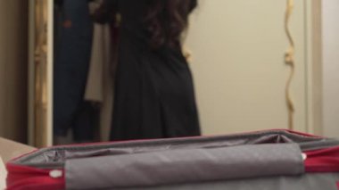 A close-up view of a red suitcase. A pretty brown-haired woman on the background opens a wardrobe, folds things, throws clothes into a suitcase and closes it.