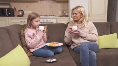 Fair-haired mum and daughter are sitting on the corner sofa and drinking tea. The girl is holding a plate with macarons on her knees. They are chatting happily