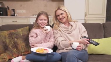 Fair-haired woman and girl are sitting on the corner sofa in the studio apartment. Woman is holding a remote control and girl is drinking tea. They are talking and laughing while watching something