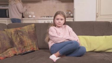 Long-haired cute girl in a knitted sweater and jeans is sitting on the corner sofa in the studio apartment and looking in front of her. Her mum is making tea in the background