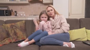 Fair-haired woman and girl sitting on the corner sofa. The girl covers face with hands, the woman strokes her head and hides her face too. The girl says something and the woman switches remote control