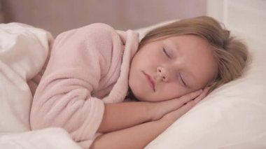 A close-up of girl's face lying sidelong and sleeping in bed. The girl opens her eyes and smiles a bit, moves her head and awakens