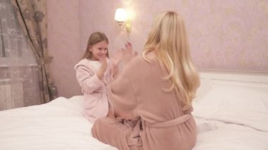 A close-up of woman and girl in gowns sitting on the bed in the bedroom and playing patty-cake. The woman is sitting back to the camera; the girl is happy and laughing