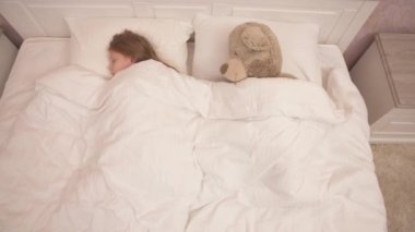 A top view of a long-haired cute girl and a big teddy bear lying sidelong on the bed under the eiderdown and covered to heads. She is sleeping