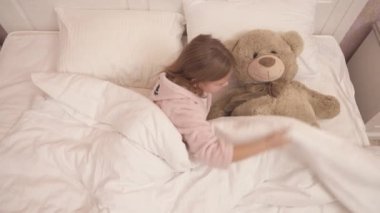 A long-haired cute girl in a gown is lying in bed and covering a big teddy bear lying beside her with eiderdown. She lies beside it and closes eyes to fall asleep
