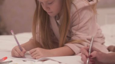 A close-up of the girl in the gown sitting on the bed, looking down and painting something with coloured pens