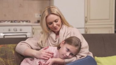 Fair-haired mum and daughter are sitting on the corner sofa in the kitchen studio. Mum is embracing and hugging the girl. They play with fingers