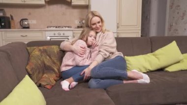 Mum and daughter are sitting on the corner sofa in the studio apartment. The woman is caressing the girl while talking and chatting