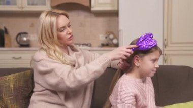 A close-up side view of fair-haired mum and daughter sitting on the corner sofa in the studio apartment. Mum is holding girl's hair and combing it before making a braid