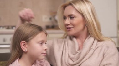 A close-up of fair-haired mum and daughter sitting and mum saying something, stroking girl's head. They touch each other's noses with pointer fingers