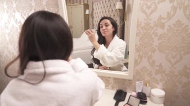 A close-up of a pretty brown-haired woman in a white gown sitting back to the camera in front of the mirror. She applies lash mascara. Woman reflection in the mirror.