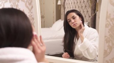 Young brunette girl reflection in the mirror. Sitting back to the camera. Holding a cotton powder puff and finishes her face make-up foundation touching her face with a sponge
