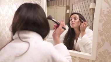 A brown-haired woman in white gown sitting in front of the mirror and holding a box with face powder. Applying powder on face: forehead, cheeks, along jaw line with a brush in light strokes.