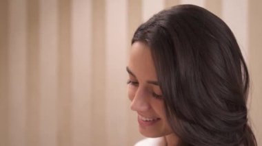 A close-up side view of young brunette's portrait. Applying face powder with a brush on cheeks, forehead, stops, turns her head a bit to check, then keeps on applying the powder.