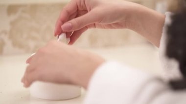 A close up of woman's manicured hands and a jar with cream lotion. Right hand twists the lid, opens the jar, takes some cream and starts applying it. Hands move slowly to spread and soak the cream