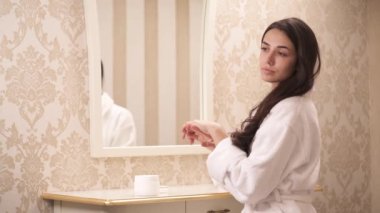 A side view of a lovely brunette with long hair in a white gown sitting in front of the mirror. She rubs some cream into hands looking to the left pensively. Looking at the camera and smiling