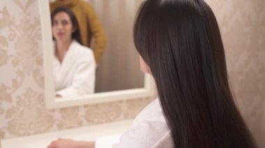 A charming long-haired brunette sits back to the camera saying something. A woman's hand brushes her hair to the length and untwines one hair strand