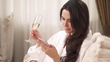 A close up of a charming brunette holding a phone and looking at the screen. She holds a wine glass and scrolls a phone