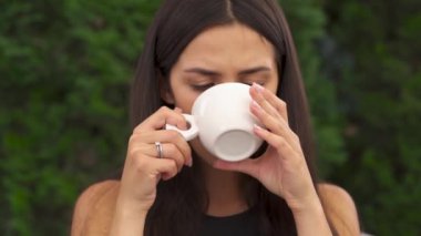 A close-up of young brunette's woman sipping coffee slowly. She tastes coffee, nods head and keeps on drinking