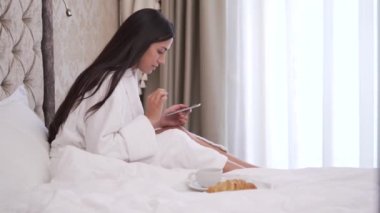 A young brunette is sitting on bed looking at the phone, drinking coffee or tea. She puts the cup back and types something.