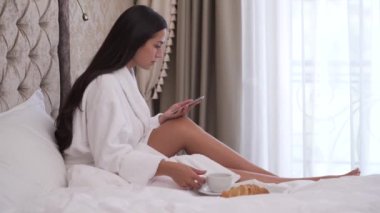 A close-up sideview of a young brunette sitting on bed and scrolling a phone. She drinks tea or coffee, smiles and continues reading and typing.