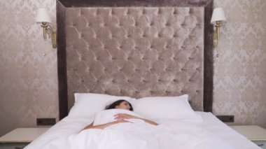 A young brunette is covered with white blanket and is slowly awakening. She opens her eyes, turns her head, then sits, looks around, stretches a bit and falls back pulling the blanket over her head.