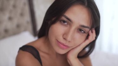 A portrait of a young brunette sitting in the bedroom with pensive look. The camera moves away a bit.