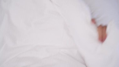A top view of young brunette's face peeping out the blanket. She smiles, looks into the camera, hides behind the blanket again.
