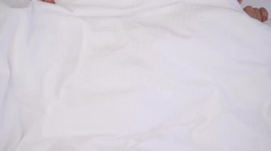 A top view of young brunette's eyes slowly peeping out the blanket. She looks into the camera, blinks, rotates eyes to the nose, hides under the blanket again.
