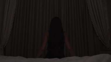 A long-haired brunette sits on the bed facing the closed window in the dark room. She stands up, goes to the window, opens the curtains. She stretches her hands and moves the body.