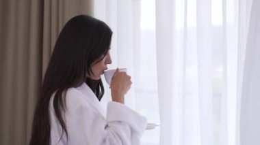 A close up of a young brunette in profile standing in front of the window. She is holding a cup and a saucer, makes one sip, starts laughing and turns head to look into the camera.