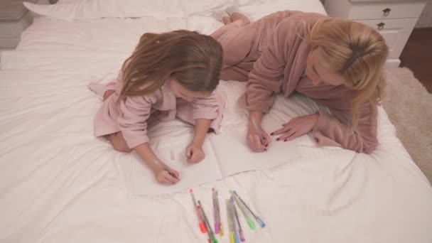 ベッドの上に寝そべってノートに色のついたペンで何かを描くガウンを着た母親と娘のトップビュー — ストック動画