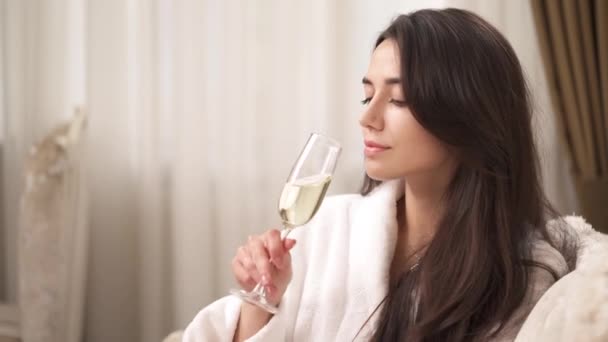 白いガウンを着た魅力的なブルネットの側面図が部屋に座っています 彼女は満杯のワイングラスを取り 一口を作り 彼女の手の上に頭を傾け 彼女の前で貫通的に見えます — ストック動画
