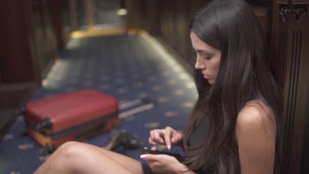 旅馆走廊的地板上坐着一个漂亮的黑头发姑娘 她靠在墙上 手提箱在地板上 她卷起电话 把它收起来 悲伤地低下了头 — 图库视频影像