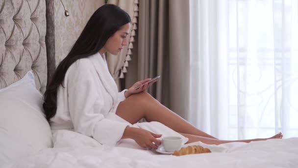 Close Sideview Young Brunette Sitting Bed Scrolling Phone She Drinks — kuvapankkivideo