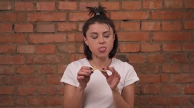 Portrait of a young funny girl in white t-shirt isolated against a brick wall background. The girl eats a delicious crispy cookie and chews it.