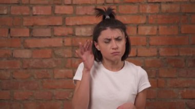 Close-up view of a funny girl in white t-shirt isolated against a brick wall background. Something got the girl in the eyes and her face twisted because of unpleasant feeling.