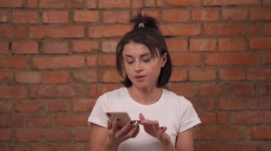Close-up view of a young girl isolated against a brick wall background. The woman uses a mobile phone and looks with uncertainty into the camera.