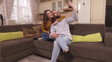 A young couple is sitting on the corner sofa and is making a selfie, turn to another side and pull faces for a selfie