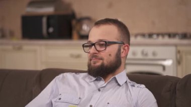 A bearded man in glasses sitting on the sofa. A brunette comes to him and hugs around his neck. He kisses her on cheek and says something to her ear. She bends above his face covering it with hair