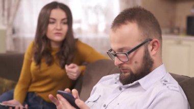 A close up of a young brunette and a bearded man in glasses sitting on the sofa with phones. She says something to him; he nods his head scrolling the phone and answers