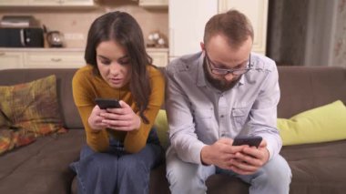 A young brunette and bearded man in glasses are sitting on the corner sofa in the studio apartment holding phones. She is typing and saying something. He is looking at the screen and scrolling it